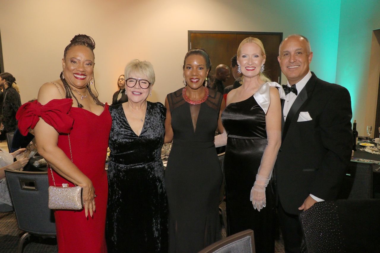 Guests in black-tie evening wear at a San Bernardino Symphony gala — women in floor-length gowns, one in a red gown with white opera gloves, beside a man in a tuxedo